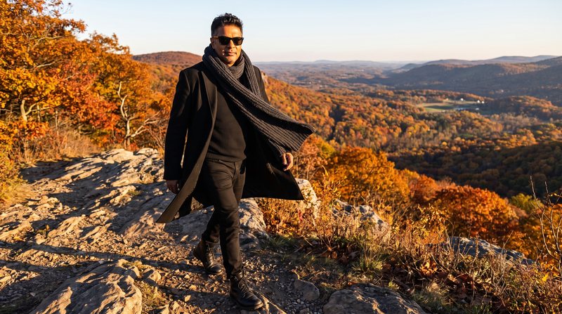 Richie Etwaru striding a ridge trail in a long black coat and oversized scarf